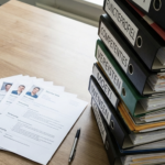 Op de afbeelding zie je een enorme stapel met mappen met daarin de eisen die de organisatie stelt aan de gezochte privacy expert. Daarnaast ligt een heel dun stapeltje met vier CV's van mogelijke kandidaten.